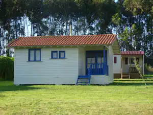 Weißer Bungalow mit rotem Ziegeldach und blauer Veranda, davor Rasen und Bäume im Hintergrund.