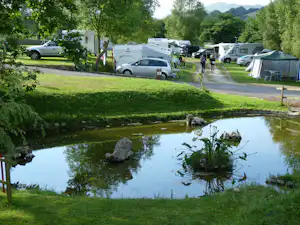 Campingplatz mit Wohnwagen und Zelten, gepflastertem Weg und kleinem Teich im Vordergrund, umgeben von Bäumen und Rasen.