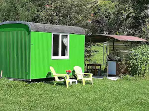 Grüner Wohnwagen mit Fenster und Eingang, davor weiße Gartenstühle und Tisch auf einer kleinen Terrasse, umgeben von Büschen.