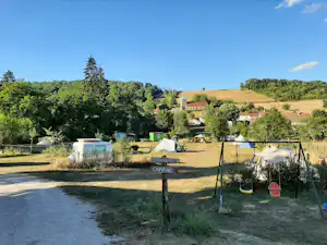 Campingplatz mit mehreren Zelten, einem Wohnwagen und einem grünen Zaun, im Hintergrund Felder und Häuser im Tal.