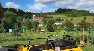 Gelbes Campingfahrzeug mit Spielplatzschaukel im Vordergrund, dahinter Wiesen, Bäume und Gebäude auf dem Campinggelände.