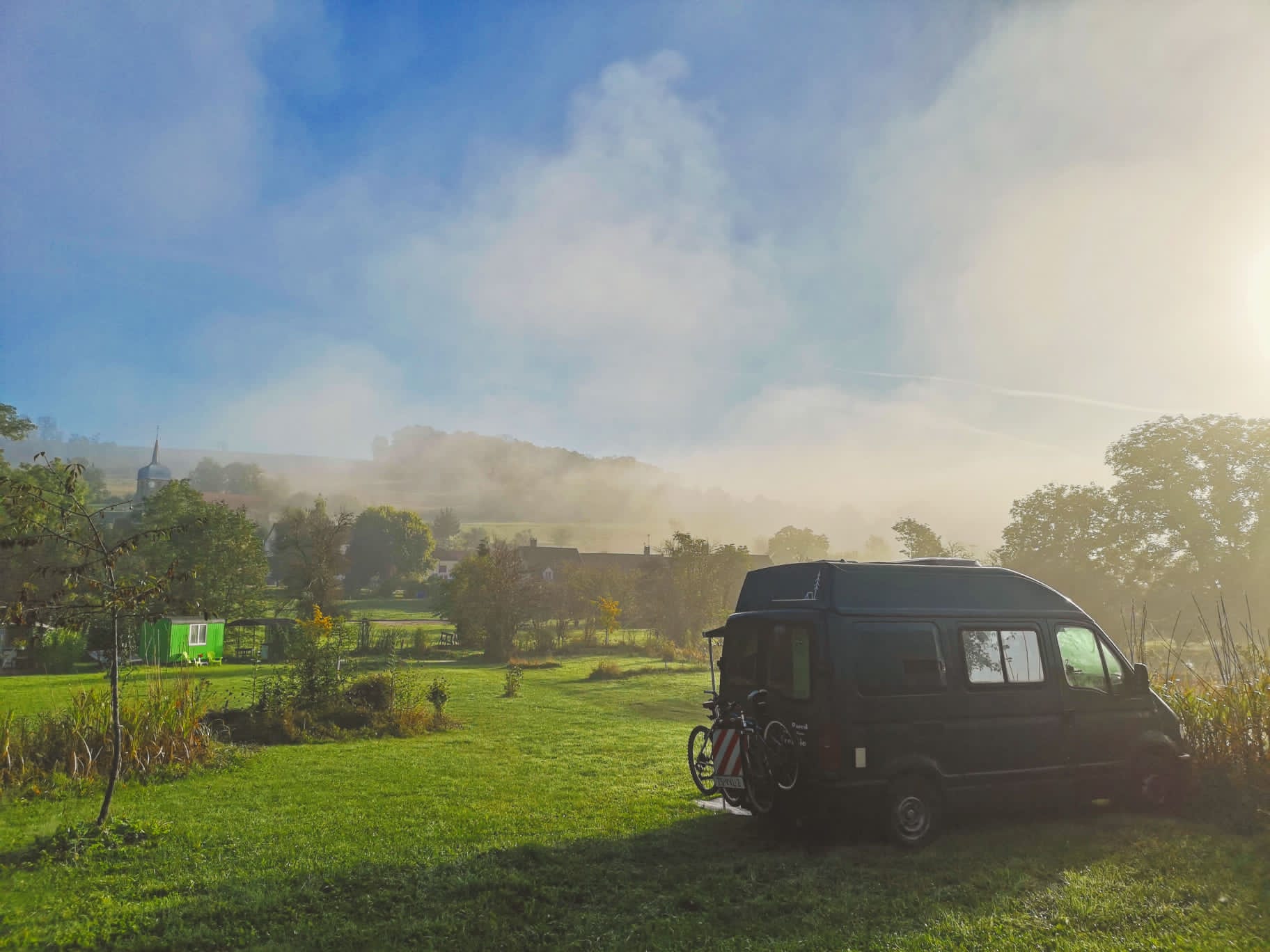 Camping-Van steht auf einer grünen Wiese am Rand eines Feldwegs, im Hintergrund Bäume und ein grünes Landschaftspanorama.