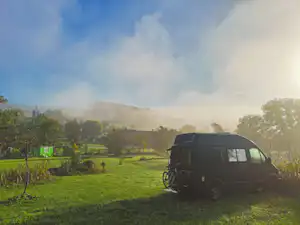 Camping-Van steht auf einer grünen Wiese am Rand eines Feldwegs, im Hintergrund Bäume und ein grünes Landschaftspanorama.