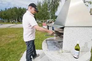 Mann am Grill im Campingbereich, Steingrill mit Flasche und Glas, Rasen und Bäume im Hintergrund.