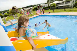 Kinder im Camping-Pool auf bunten Schwimmflößen, im Hintergrund Wohnwagen und Rasen mit Hecken.
