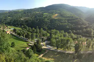 Blick von oben auf einen Campingplatz mit Wohnhäusern, Zufahrtsstraße, Stellplätzen und Tennisplatz inmitten grüner Hügel und Wälder.