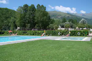 Schwimmbecken auf dem Campingplatz mit Liegestühlen, Hecken und Spielgeräten, im Hintergrund Bäume und Berge.
