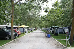 Campingplatz-Gasse mit Wohnwagen und Zelten, Autos am Rand, Bäume spenden Schatten und ein Kiesweg führt nach vorn.