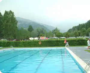 Camping-Pool mit Liegefläche, im Hintergrund Zelte und Wohnwagen sowie grüne Bäume und Berge.
