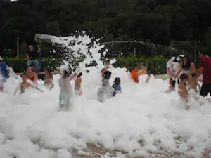 Kinder und Erwachsene feiern eine Schaum-Party im Campingpool, umgeben von Bäumen und einem Zaun im Hintergrund.