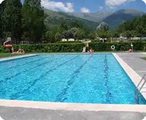 Blauer Swimmingpool mit Rand und Liegen, im Hintergrund Bäume und Berge, dahinter ein Campingplatz mit Wegen.
