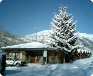Schneebedeckter Campingplatz mit Holzhütte, Pavillon und geparktem Auto, im Hintergrund Tannen und verschneite Berge.