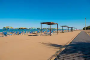 Strandpromenade mit Liegestühlen und Sonnenschirmen, im Hintergrund das Meer und ein Holzsteg mit Geländern.