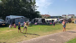 Zeltbereich auf dem Campingplatz: Kinder spielen auf dem Rasen, im Hintergrund Wohnwagen und Zelte unter Bäumen.