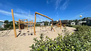 Spielplatz am Campingplatz mit Klettergerüst aus Holz, Sandboden und Bäumen sowie Wohnhäusern im Hintergrund.