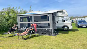 Wohnmobil auf dem Campingplatz mit ausgeklappter Markise, Sitzgruppe und weiteren Fahrzeugen auf dem Rasen.