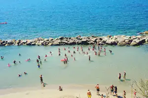 Badebucht am Meer mit vielen Schwimmern im flachen Wasser, Sandstrand und Steinschutzdamm im Hintergrund.