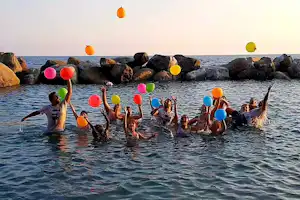 Kinder im Meer mit bunten Luftballons, im Hintergrund Felsen und Küste bei ruhiger Wasseroberfläche.