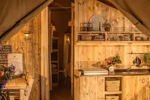 Innenansicht eines Campingzelts mit Holz-Küchenzeile, Spüle, Schränken und Regalen, gedecktem Tisch und Vorhängen.