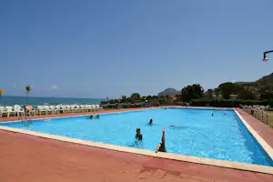 Blaues Camping-Schwimmbecken mit Liegestühlen am Rand, Blick aufs Meer und auf Bäume am Ufer.