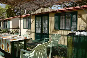 Bungalow mit grünen Türen und Fenstern, überdachte Terrasse mit Tisch, Stühlen und Sonnenschirm, im Hintergrund Bäume.