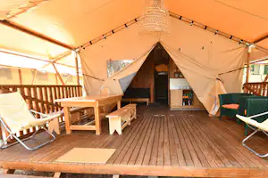Innenansicht eines Campingzelts mit Holzterrasse, Sitzbank, Stühlen und einem kleinen Küchenbereich im Hintergrund.