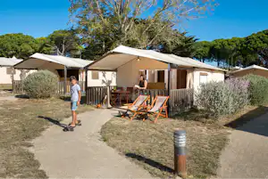 Campingzelt mit überdachter Terrasse, Holzgeländer und Liegestühlen am Weg, umgeben von Bäumen und Sträuchern.