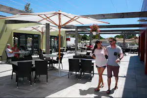 Terrasse eines Campingplatzes mit überdachtem Bereich, vielen schwarzen Tischen und Stühlen sowie zwei Personen.