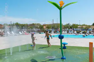 Sprühfontäne im Camping-Schwimmbecken, Kinder planschen, daneben liegen Liegestühle und Bäume vor dem Poolbereich.