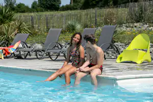 Kinder im Pool eines Campingplatzes, im Hintergrund Liegen und ein grüner Sonnenschirm, Garten mit Hecke und Blumenbeeten.