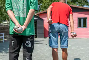 Zwei Männer spielen Boule auf einem Campingplatz, mit roten und grünen Shirts, Kugeln in der Hand und Wohngebäuden im Hintergrund.