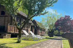 Steinweg und Rasen im Campingbereich, davor mehrere Liegestühle vor Holzhäusern, umgeben von Bäumen und Hecken.