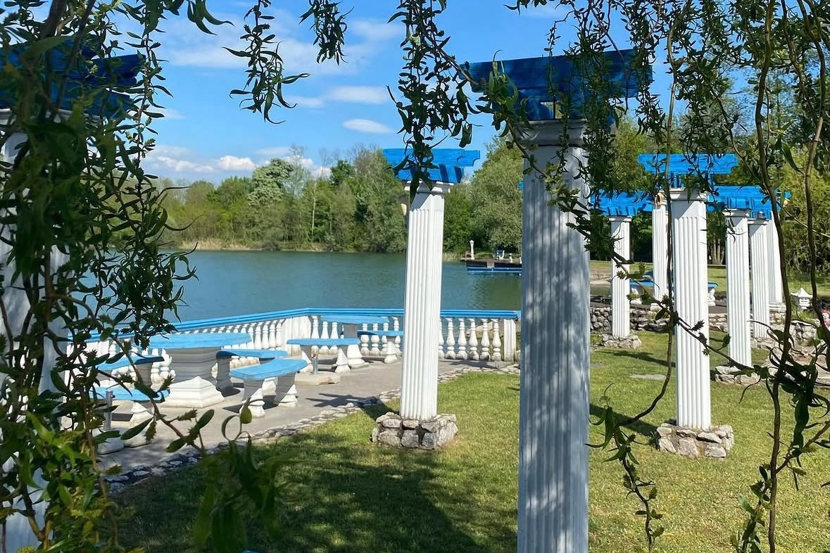 Blick auf den See mit Liegestühlen am Ufer, Rasen und weißen Säulen unter Bäumen im Campingbereich.