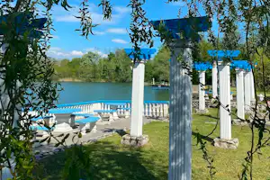 Blick auf den See mit Liegestühlen am Ufer, Rasen und weißen Säulen unter Bäumen im Campingbereich.