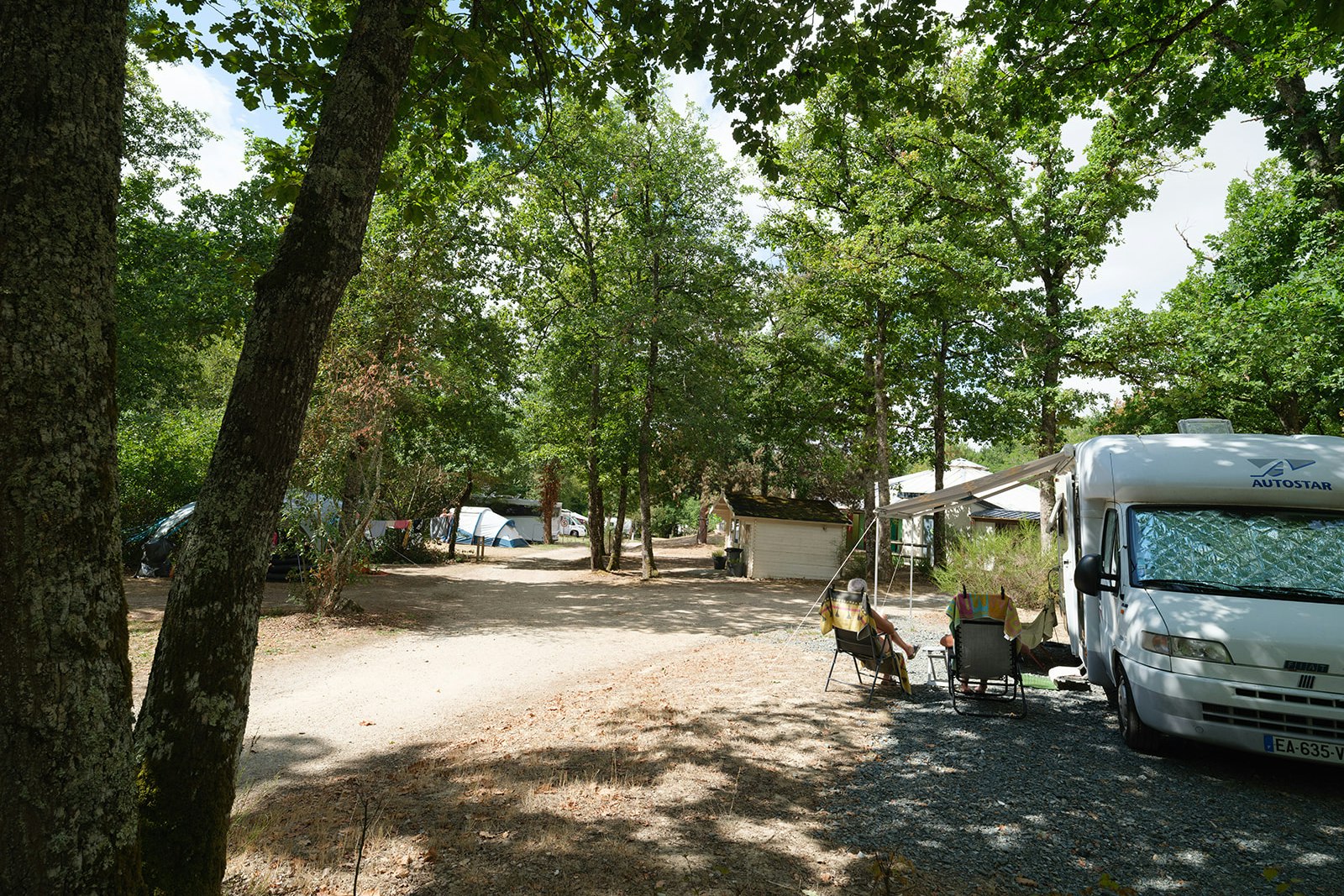 Campingplatz mit Stellplätzen im Schatten der Bäume, Wohnmobil rechts und Zelt im Hintergrund entlang des Kieswegs.