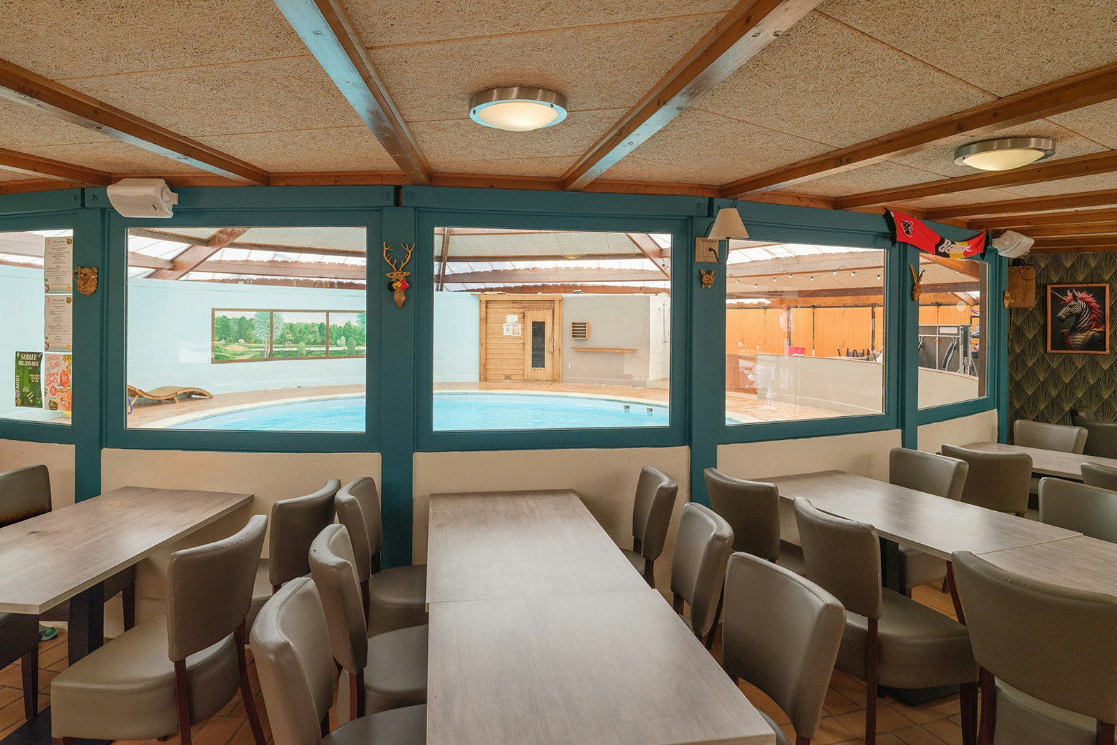 Innenbereich eines Campinghauses mit langen Tischen und Stühlen unter einer Holzdecke, große Fenster mit Blick nach draußen.