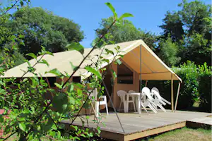 Zelt auf Holzterrasse im Grünen, weiße Campingstühle und Liegestuhl, umrahmt von Bäumen und blauem Himmel.