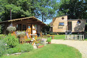 Holzhütte mit Schild „ACCUEIL“, davor Sitzbank und Blumenbeete, daneben ein Wohnwagen auf Rädern im Grünen.