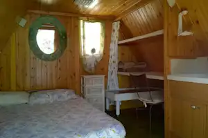 Innenraum einer Holzunterkunft mit Doppelbett, Schrank, Regalen und Fenster, Blick auf Vorhang und Spiegel.