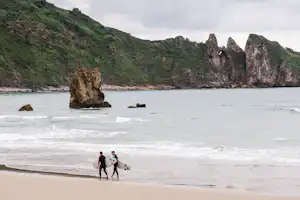 Strand mit Steilküste und Felsen im Wasser, zwei Personen laufen am Ufer entlang, Wellen rollen an.