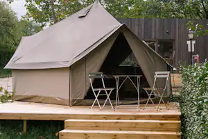 Zelt auf einem Holzpodest mit zwei Klappstühlen und kleinem Tisch, umgeben von Bäumen und grünem Rasen.