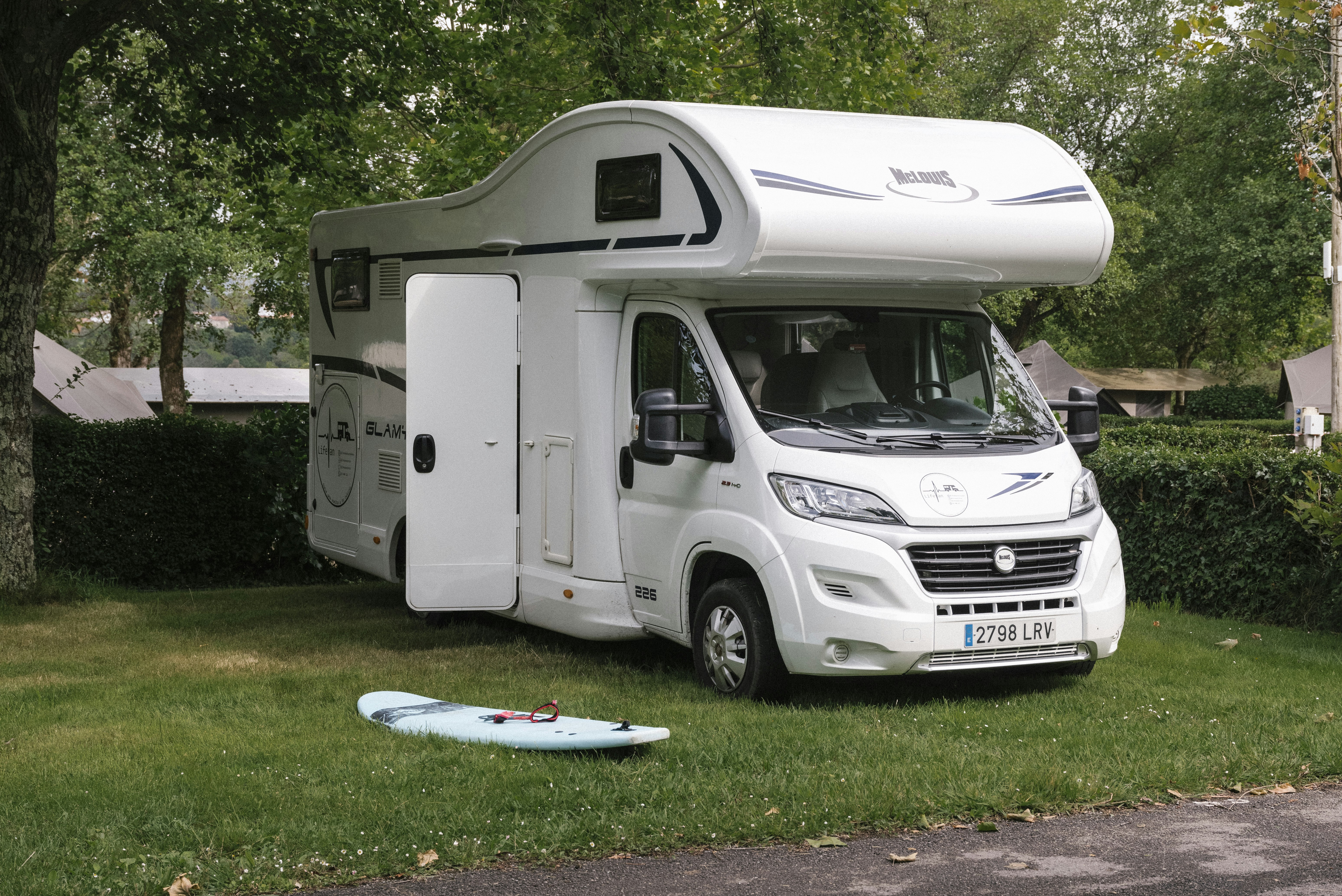 Weißes Wohnmobil auf einem Campingplatz im Grünen, mit geöffneter Markise und Blick auf Bäume und Rasen.