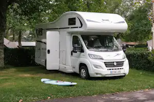 Weißes Wohnmobil auf einem Campingplatz im Grünen, mit geöffneter Markise und Blick auf Bäume und Rasen.