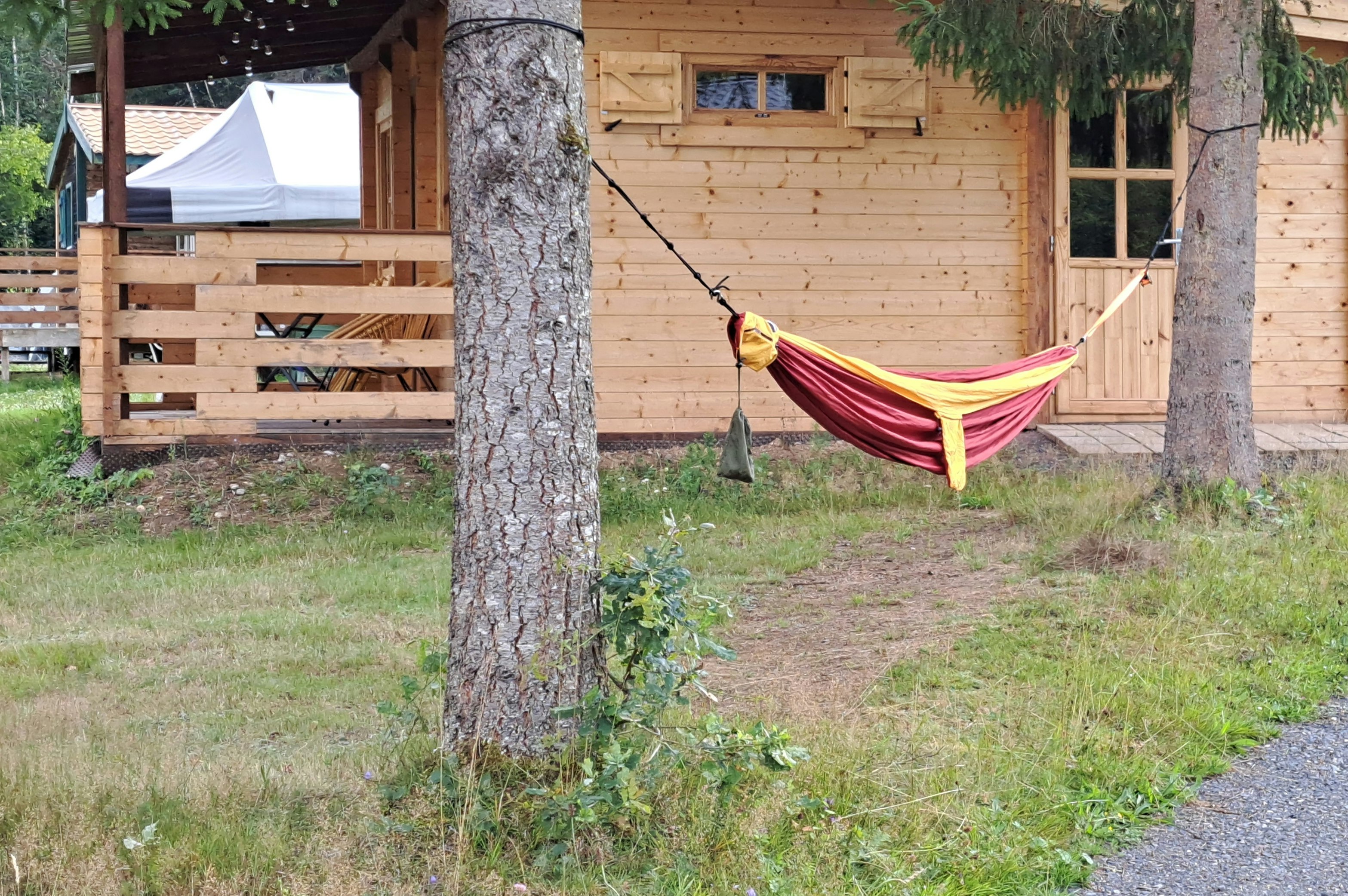 Holzhaus am Campingplatz mit Veranda, Hängematte zwischen Bäumen und Kiesweg im Garten.