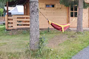 Holzhaus am Campingplatz mit Veranda, Hängematte zwischen Bäumen und Kiesweg im Garten.