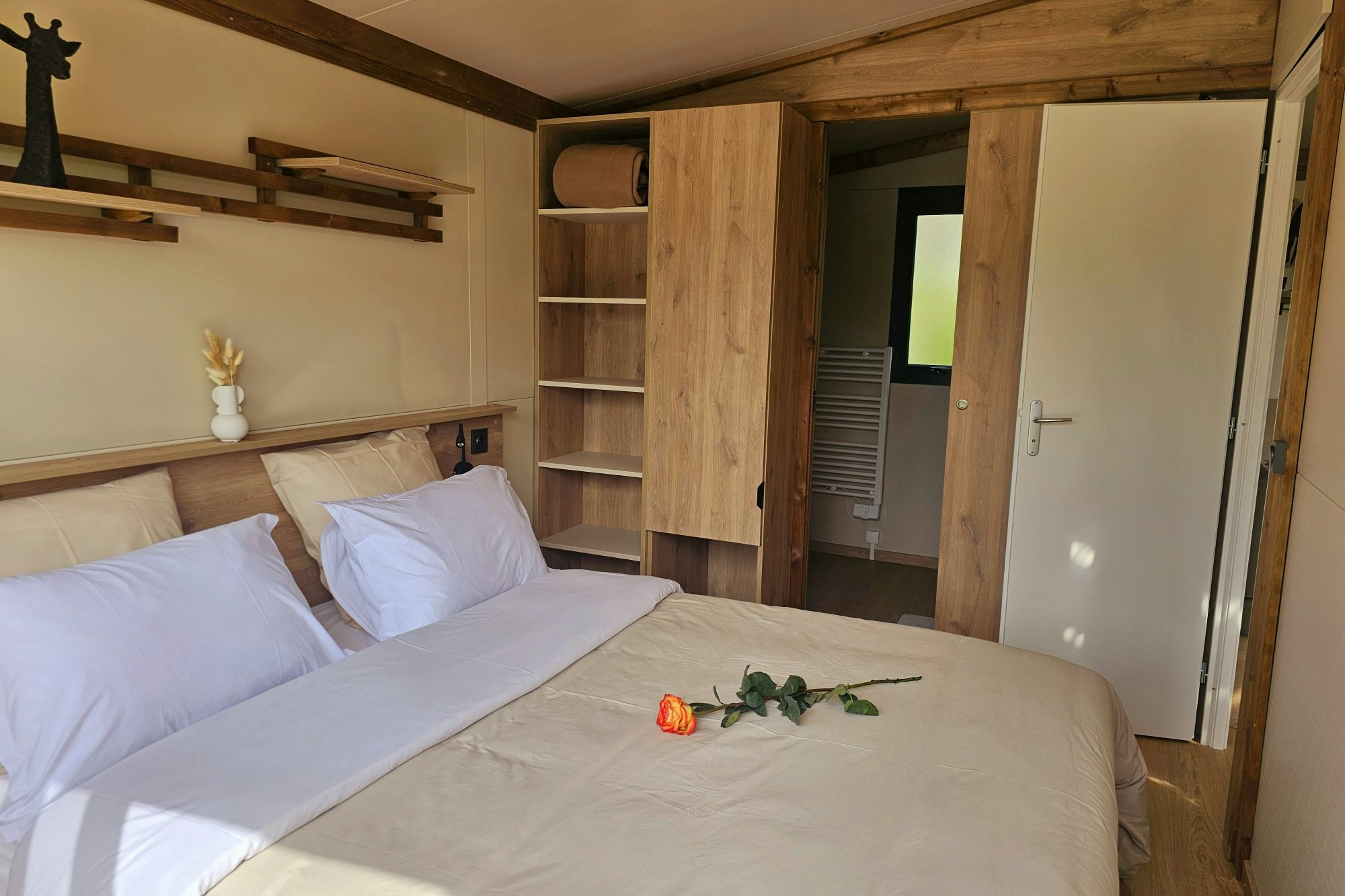 Schlafzimmer in einem Mobilheim: zwei Betten, Holzregale und Schranktüren, daneben eine offene Tür ins Zimmer.