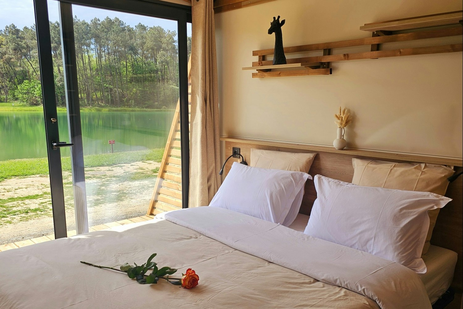 Bett in einer Campingunterkunft mit großen Fenstern und Blick auf den See, Holzdecke und Kissen auf dem Bett.