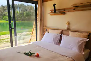 Bett in einer Campingunterkunft mit großen Fenstern und Blick auf den See, Holzdecke und Kissen auf dem Bett.