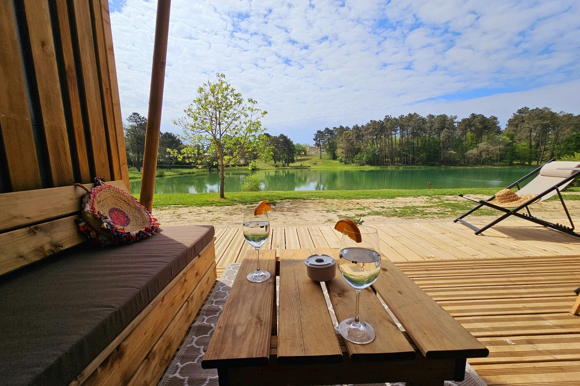 Holzterrasse am See mit Esstisch, Gläsern und zwei Liegen, im Hintergrund grünes Wasser und Bäume.