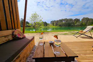 Holzterrasse am See mit Esstisch, Gläsern und zwei Liegen, im Hintergrund grünes Wasser und Bäume.
